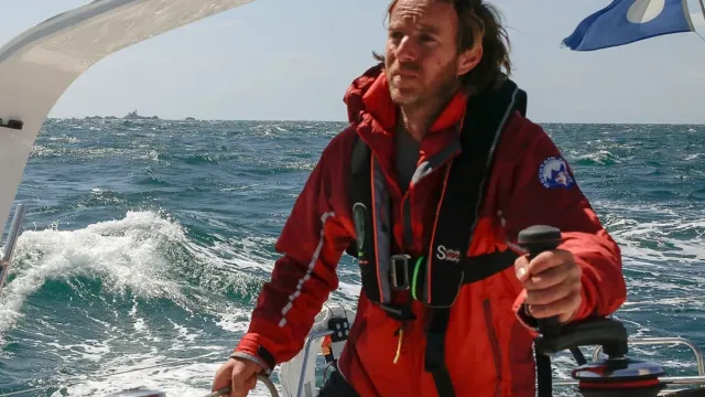 Sailing school student helming a yacht in rough seas wearing red foul-weather gear