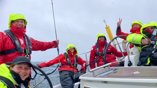 Sailing school students in red and neon foul-weather gear steering a yacht and raising a green flag in rough seas