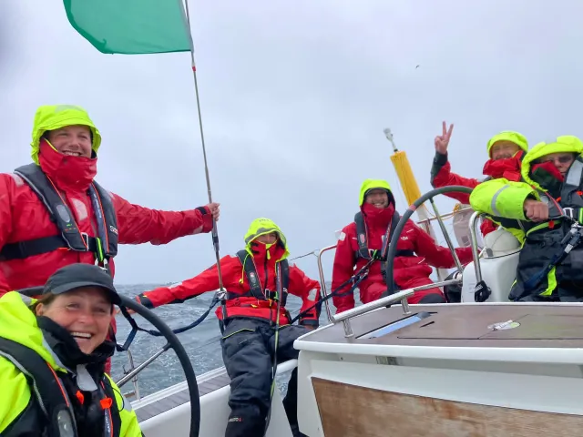Sailing school students in red and neon foul-weather gear steering a yacht and raising a green flag in rough seas