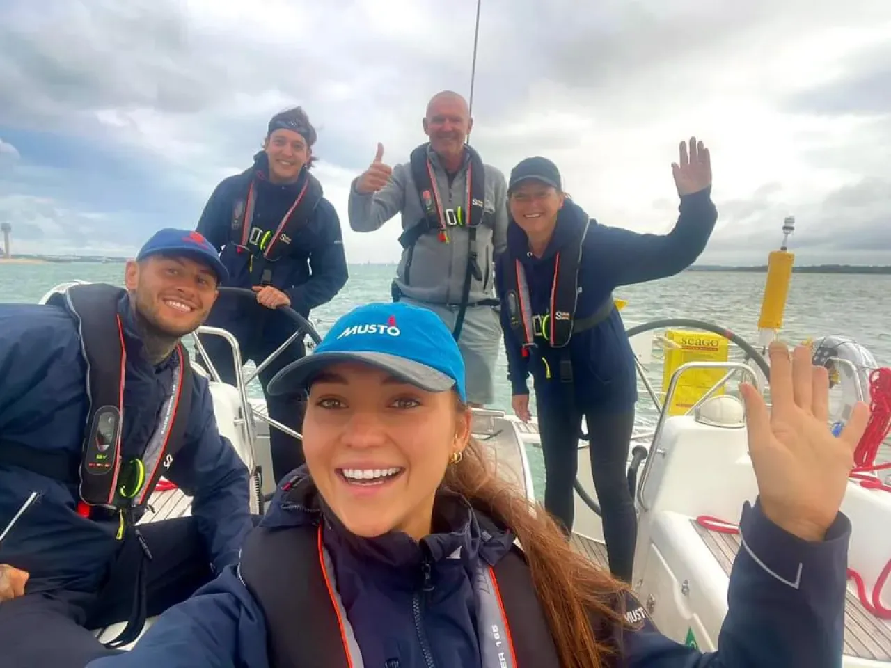 Sailing school students and instructor smiling and waving on deck during a training sail