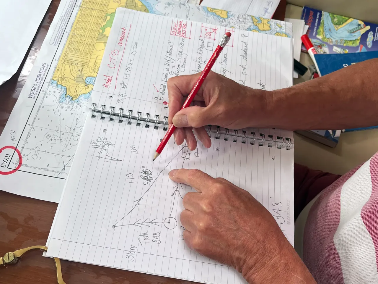 Sailing school student making navigation notes in a logbook with charts on the table